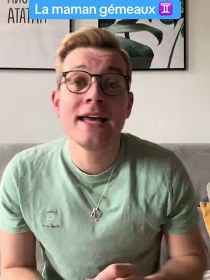 A man with glasses is centered in the frame, speaking directly to the camera. He wears a light green t-shirt and a silver necklace with a pendant. A light blue banner with white text that says, La maman gémeaux and a Gemini symbol, is at the top. The background has a neutral color scheme, including a couch and framed artwork.
