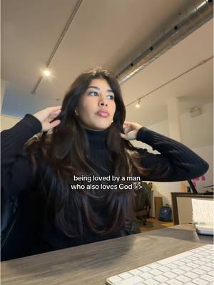 A woman with long, dark hair appears to be indoors, sitting at a desk with a keyboard in front of her. She is wearing a black turtleneck sweater and has her hands near her head as if she's adjusting her hair. The lighting is soft, suggesting an interior setting. Text is overlaid on the image.
