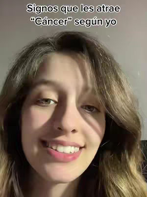 A young woman is centered in the frame, her face taking up most of the shot. She has long, wavy brown hair, and her facial expression is a slight smile with visible teeth. Above her, a white text overlay reads, "Signos que les atrae \"Cáncer\" según yo" which translates to "Signs that Cancer is attracted to, according to me". The background is blurred, suggesting an indoor setting with neutral tones.