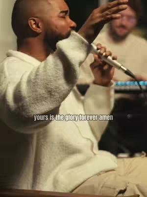 A man with a shaved head sings into a microphone, his right arm raised. Text overlayed reads, 'Yours is the glory forever amen.' Behind him, another person is visible, blurred, possibly playing a keyboard. The image has a warm, indoor lighting with a shallow depth of field.