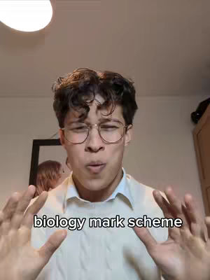 A young man with curly brown hair and glasses is centered in the frame, hands raised in front of him, with the text 'biology mark scheme' overlaid. The background is a simple room with a light fixture and a framed picture visible. The overall tone is informational.
