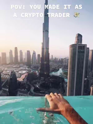 A first-person perspective of a hand reaching towards the camera with a backdrop of a city skyline featuring the Burj Khalifa skyscraper. The foreground shows turquoise water, suggesting a pool or elevated water body. The sky is a gradient of light blue. The image is composed to highlight the city view, framed by the hand in the foreground.