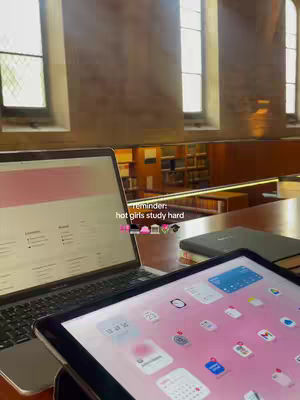 A laptop and a tablet rest on a wooden table, with a window in the background. The tablet shows a pink interface with colorful app icons. The laptop screen has a pink and white theme. Text overlaid reads "reminder: hot girls study hard."