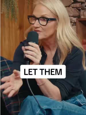A woman with blonde hair and glasses is holding a microphone and speaking. Text overlayed on top of the image says 'LET THEM.' She is wearing a dark shirt and jeans and appears to be in a studio setting.