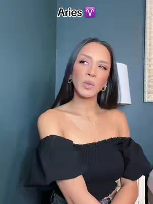 A medium shot shows a woman facing the camera against a blue-gray backdrop. She has long, dark hair and is wearing a black off-the-shoulder top and small gold hoop earrings. Text that reads "Aries" is on the screen.
