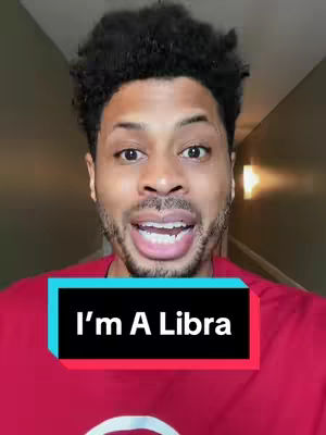 A man with short, dark, curly hair is centered in the frame, his face expressing excitement and a wide smile. He's wearing a red shirt. Below him, a graphic says "I'm A Libra" in white text inside a rectangular box with a black background and a red and blue outline. The background is a hallway with a brown door and muted lighting.