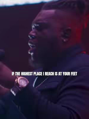 A man with a short haircut, wearing a black jacket and a watch, sings into a microphone in a dimly lit setting with a purple-toned background. Text overlayed says, "IF THE HIGHEST PLACE I REACH IS AT YOUR FEET".