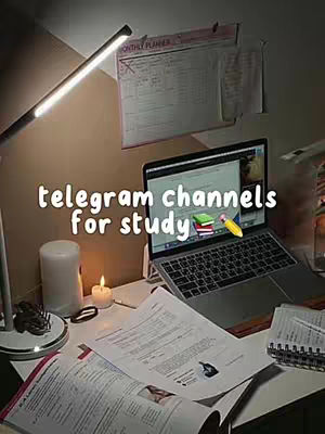 A well-lit workspace featuring a laptop displaying a website, with books, papers, a candle, and a desk lamp. The background includes a poster with a monthly planner.