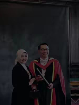 A photo shows a man and a woman, both smiling for the camera. The man wears a graduation gown with a red and black design and holds what appears to be a diploma. The woman next to him wears a black suit and a light-colored hijab. They stand in front of a dark background.