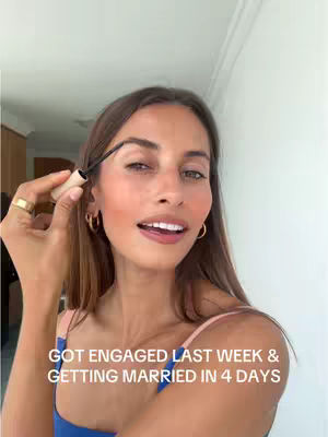 A woman is applying makeup, holding a brow product. She is smiling and looking at the camera. Text overlay reads: GOT ENGAGED LAST WEEK & GETTING MARRIED IN 4 DAYS