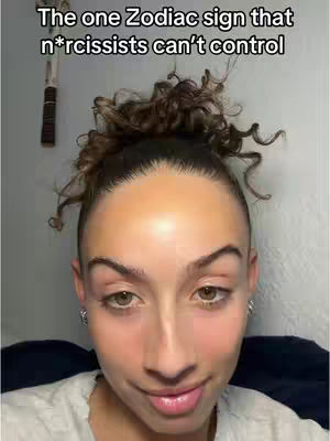A close-up shot of a woman looking directly at the camera; the woman's face is centered in the frame with the top of her head and part of the background visible; she has light skin, fair complexion with visible makeup, her eyebrows are well-defined, and her dark brown eyes are focused with a slight smirk, the background is a plain wall, and there is a small part of an unknown object visible.