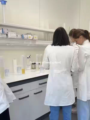 The image shows a laboratory setting with several people in white lab coats. The camera angle is from behind the group, focusing on their backs as they work at a counter with various beakers and containers. Shelves with boxes and equipment are visible in the background adding to the scientific environment.