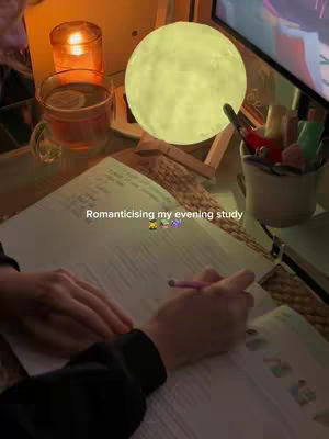 The image shows a person studying in a cozy, well-lit setting. A soft, round moon-shaped lamp casts a warm yellow glow, complemented by a lit candle in a small glass. A cup of tea rests on the table, beside an open book. The hands of the person are visible, holding a pencil and writing on the pages. The scene is bathed in a warm, inviting light, creating a comfortable atmosphere reminiscent of a relaxing evening spent studying.