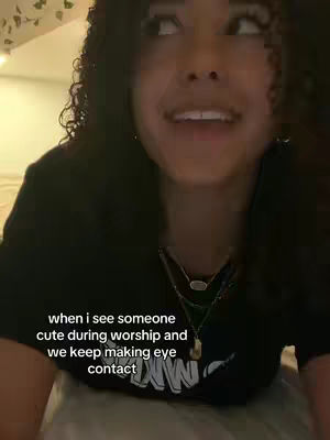 A woman with dark, curly hair is looking up and slightly to the right, smiling. The background is softly lit, with a pattern visible, and the woman is wearing a dark shirt. Text overlayed at the bottom reads, “when i see someone cute during worship and we keep making eye contact”.