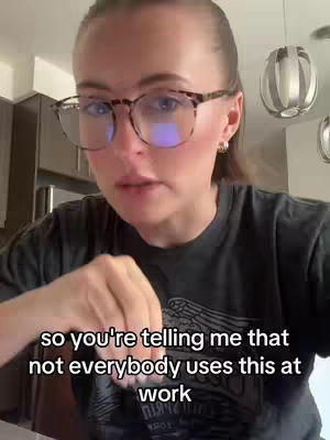 A woman with fair skin and light-brown hair, wearing glasses and earrings, looks directly at the camera. She is wearing a dark t-shirt. Kitchen cabinets and lights are visible in the background.