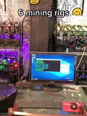 The image shows a close-up view of a crypto mining setup. The foreground features a desk with a keyboard, mouse, and a small digital display. A computer monitor displays what seems to be mining software. Behind the desk, there are multiple mining rigs stacked with hardware. The background is filled with various components, including fans and electrical equipment, which provides context about the video content. The image uses a color palette mainly of blues, greens, purples, and black.
