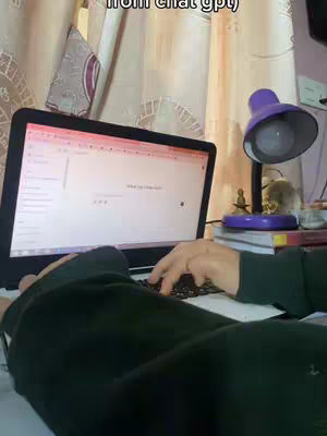 A person's hands are typing on a laptop's keyboard, with a laptop displaying a webpage. The lighting is soft and the background has a purple lamp and other objects.