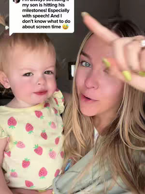 A woman points her finger at the camera while holding a small child wearing a strawberry-patterned top.