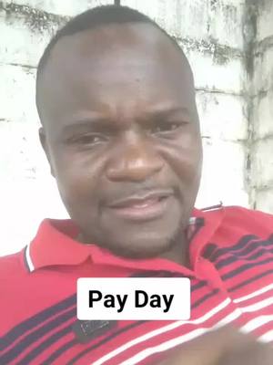 A man with short black hair is centered in the frame, looking directly at the viewer with a serious expression. He's wearing a red polo shirt with white stripes on the collar. Below him, a white rectangle reads 'Pay Day' in a bold, black font. The background is a plain wall, slightly out of focus.