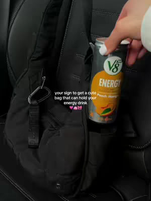 A vertical shot showing a partially visible black bag with a silver carabiner clip hanging from it. A hand holding a V8 Energy drink can is visible, with text overlayed on the image.