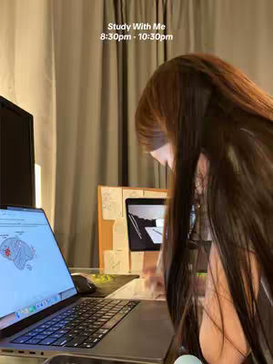 A person is seated at a desk, with a laptop open in front of them. The screen displays a diagram. The person's long hair is partially obscuring their face, they are in front of curtains, and the scene is illuminated by ambient light.