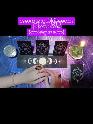 The cover image showcases a tarot card reading setup. Three tarot cards are arranged in a row above a decorative wooden block featuring lunar and celestial symbols. Below, the hands of a person are visible, adorned with rings and a bracelet, interacting with the cards. The background is a patterned cloth in shades of purple, with additional tarot cards and candles placed to the sides, creating an overall mystical and spiritual ambiance.