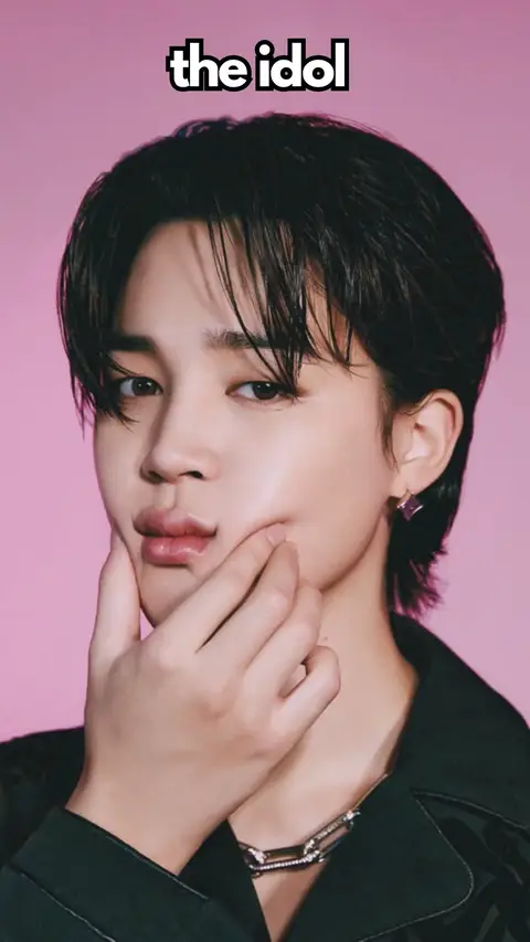 A close-up shot featuring a light-skinned man with dark hair and a pink background. He is looking towards the camera with his index finger touching his full lips. The text "the idol" is overlaid above. He’s wearing a dark, possibly black, jacket and a silver chain necklace.