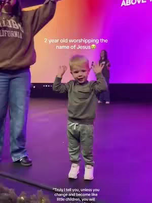 The image captures a vibrant indoor scene during what appears to be a church service or worship event. The focus is on a young, fair-skinned boy, probably around two years old, who is standing in the center of the frame. His arms are raised in the air, and he has a joyful expression on his face, possibly indicating he is participating in a worship song or activity. The background includes several figures, likely other worshipers, and a colorful backdrop of vibrant colors. The lighting appears to be the colors of a stage. Adding text overlay. The boy is wearing casual clothes, and his attire complements the candid and spontaneous mood of the scene.