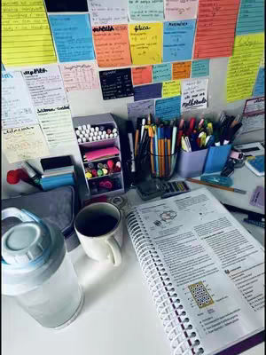 A well-organized desk with study materials. A notebook is open with detailed notes, surrounded by pens, markers, a cup of coffee, a water bottle, and sticky notes on the wall.