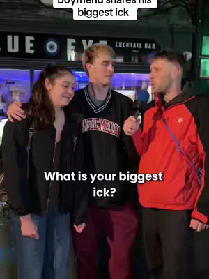 An image capturing a street interview scene with three individuals. In the center, a young man and a woman stand close together while the interviewer, a man with a microphone, stands at their side. The background features the exterior of a bar with a dark ambiance, providing a glimpse into the setting of the interaction.