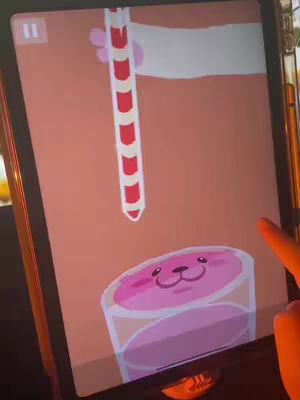 A digital illustration on the screen of a tablet shows gameplay. A cartoon hand is holding a striped tube with a pink liquid underneath. Below, a glass contains pink fluid with a smiling face on top. The background is a solid peach color.
