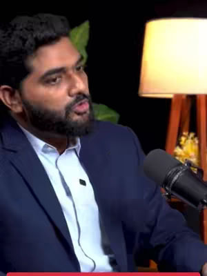 A man in a navy suit sits in front of a dark background with a lamp in the background. He is holding a microphone. The lighting seems to be coming from in front of him. The background seems to be a dark curtain.