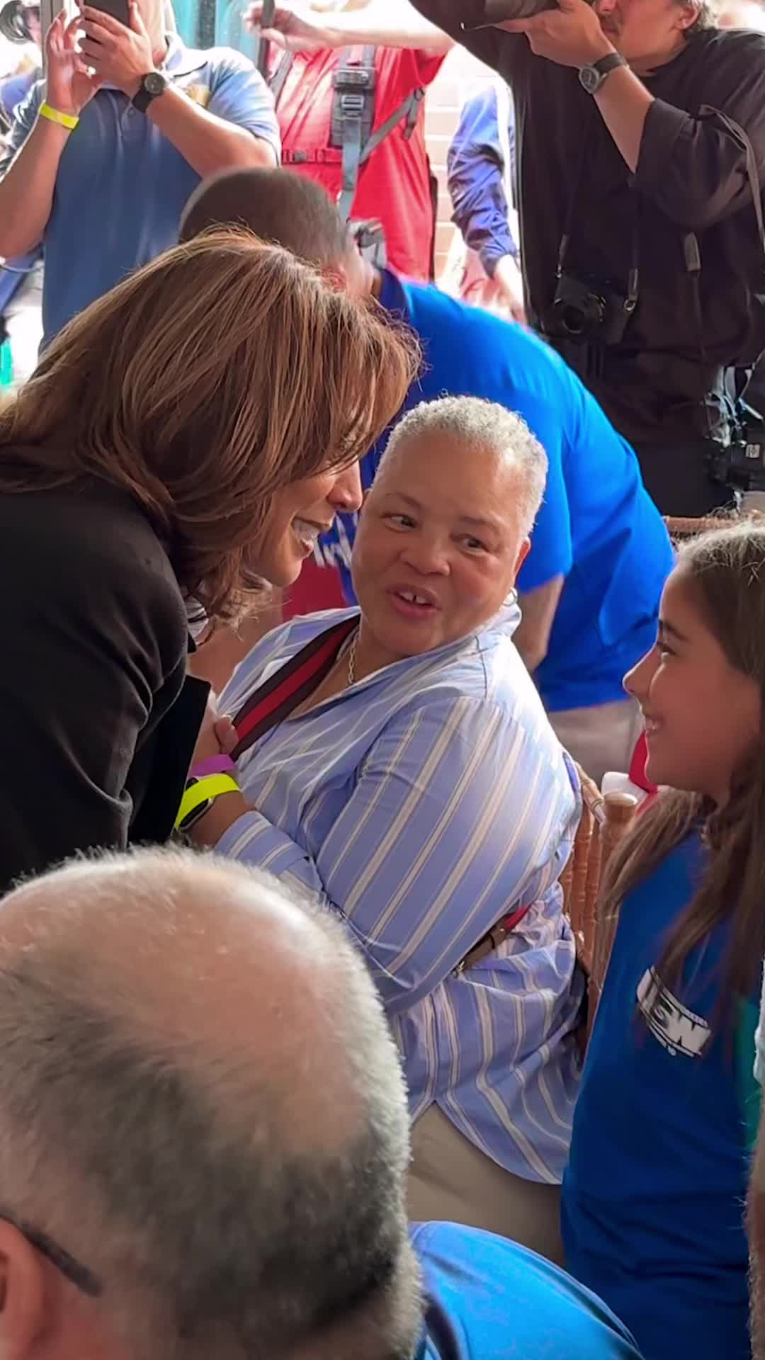 An image of three people. One is in the center, with short hair and a light blue striped shirt, looking towards the person to the right. The person on the right is a young girl, wearing a blue shirt. The person on the left is facing the person in the middle with short brown hair and a black blazer over a white shirt. Several other people are in the background.
