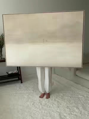 A person in white pants and pink bare feet holds a large framed artwork in front of their face. The art is a light neutral color with a horizon line in the center and two small figures. The person is standing on a white fluffy rug.