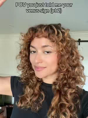 A woman with curly brown hair and a black top is centered in the frame, facing the camera. Text in white at the top reads \"POV you just told me your venus sign (pt 2)\". The background is blurry, with hints of a room. The lighting is bright, and the overall composition focuses on the woman's expression.