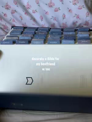 A close-up shot of a Bible with tabs labeled with names of the books, with the text overlay "decorate a Bible for my bestfriend w/me". The bible is covered with a white fabric, and behind it, a flowery pattern. The overall composition is neat and organized with a soft color palette.