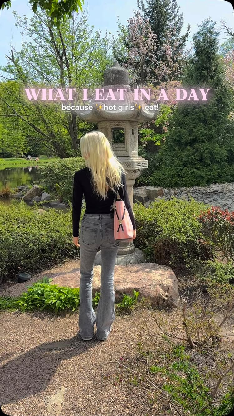 A woman stands in a garden, facing away from the camera. She wears a black long-sleeved shirt and gray jeans, with blonde hair that cascades down her back. She carries a pink purse. The background includes lush greenery, trees, a garden structure, and a pond.