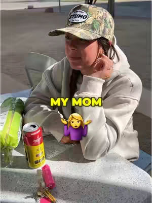 A selfie-style shot shows a person wearing a camouflage baseball cap and a beige hoodie. The person appears to be outside, with a can of soda and other items in front of them on a white table. Yellow text on the image says 'My Mom,' with an emoji of a blonde woman with her arms raised.