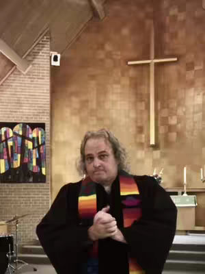 A man in a black robe and colorful stole stands in a church, hands clasped in front of him. A cross hangs on the wall behind him. A piece of abstract art is visible.