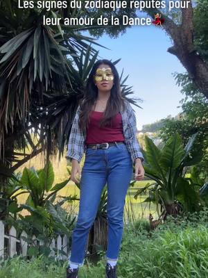 A woman stands confidently amidst lush greenery and a blue sky, wearing a gold mask, blue jeans, and a red top. The image has a vibrant and natural feel, with a mix of sunlight and shadows.