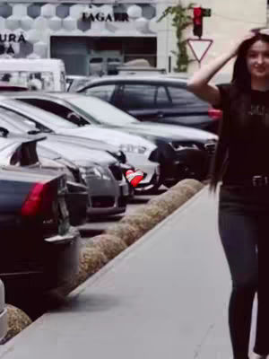 A woman standing on a sidewalk with cars parked on the side. She is wearing black. The background features a building and street details.