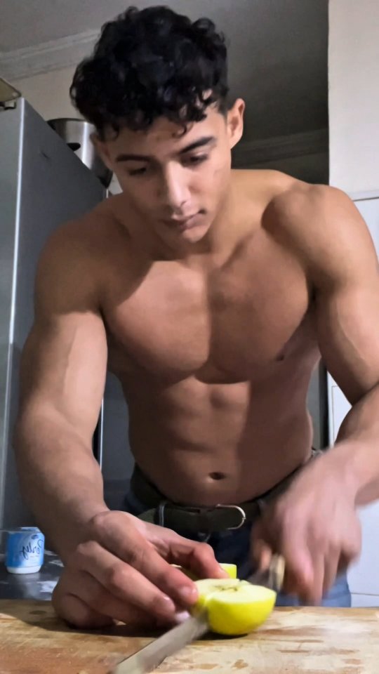 A muscular man with dark curly hair is shown shirtless from the chest up, preparing food in a kitchen setting. He is holding a knife and cutting a yellow apple on a wooden cutting board. The background includes a stainless steel appliance and kitchen cabinets.