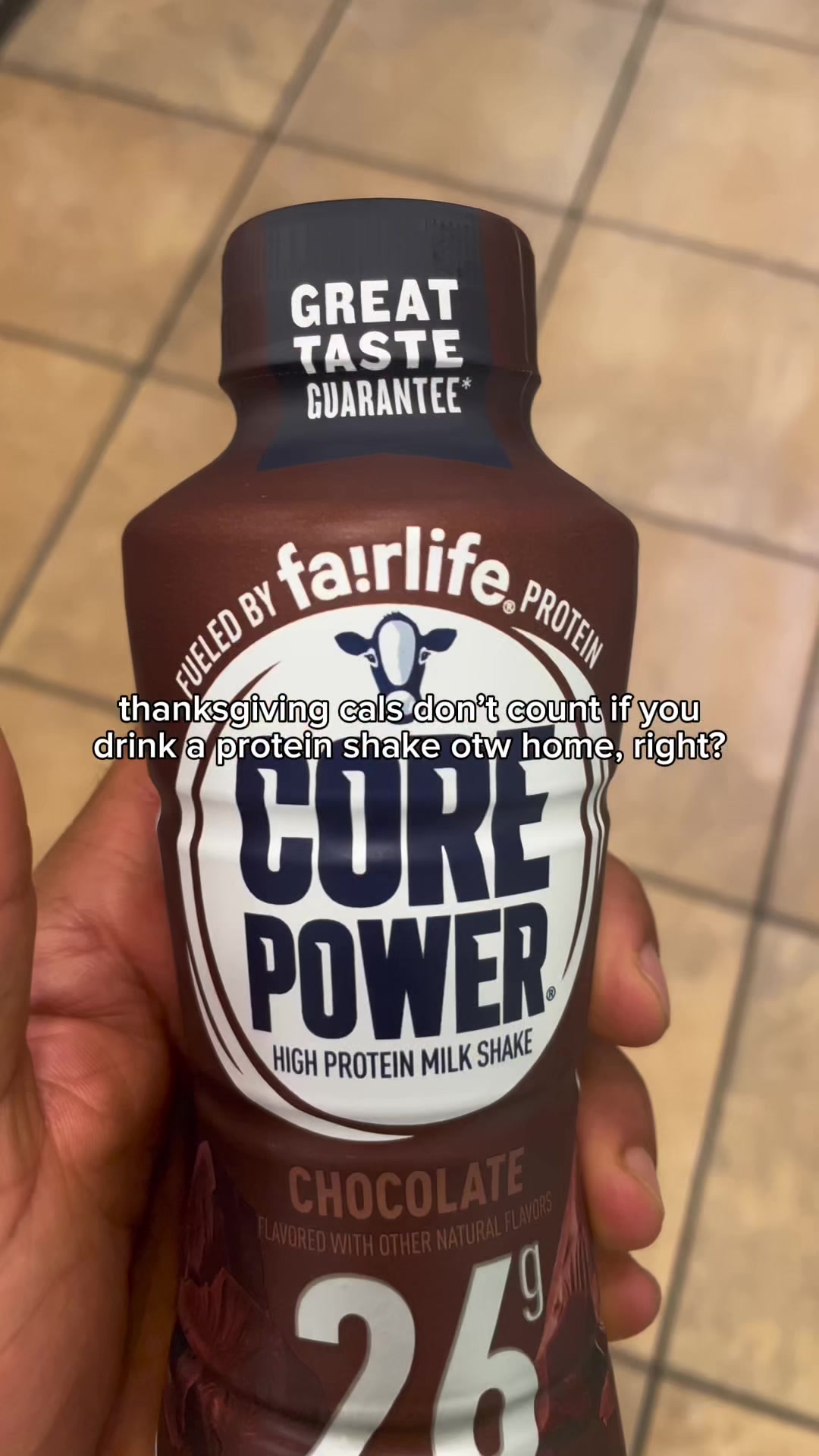 The image shows a close-up of a chocolate-flavored Core Power protein shake bottle held in a hand. The bottle has a white and blue label with text, and the background is a light brown, possibly tiled surface.