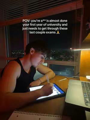 A young man studies at night, illuminated by a laptop screen and a desk lamp, with a city view outside the window.