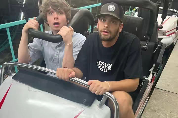 Two young men are on a roller coaster, seated side-by-side in a car that appears to be in motion. The man on the left has wide eyes and a look of surprise, holding onto the safety bar. The man on the right has a more composed, yet slightly anxious, look. He's wearing a black t-shirt with 'JESUS LOVES YOU' in white letters. The background is blurred, suggesting speed or motion. They are the central focus, and their expressions are key to the image.