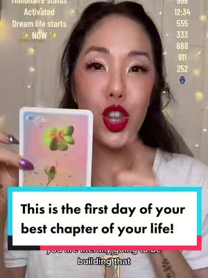 A selfie-style video features a woman holding a tarot card in the foreground. She has dark hair, makeup is present, and her lips are a vibrant red. A hook at the bottom of the image is visible as well with another text on the top left side of the image. Behind the woman, there is a warm-toned background with a blurred effect, adorned with subtle fairy lights enhancing the ambiance.
