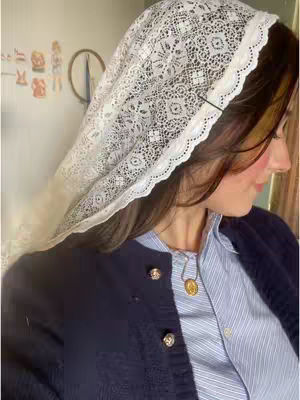 A woman with brown hair is seen in profile, wearing a white lace veil and a navy blue cardigan over a light blue striped shirt. She has rosy cheeks and a gold necklace with a medal. The background is a softly lit room with a subtle wall design.