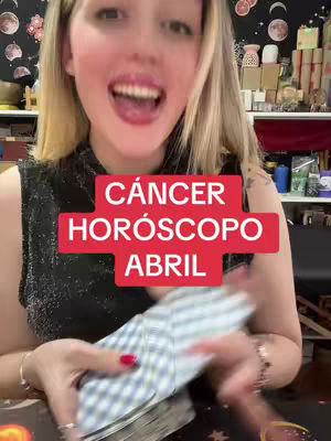 A woman with long blonde hair, smiling widely with bright red lipstick. She is holding a deck of cards and has a red rectangle overlay with white text that reads "CÁNCER HORÓSCOPO ABRIL". The image is well-lit, and the background is slightly blurred with shelves and items.