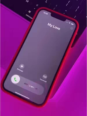 An iPhone on a purple lit surface, likely a desk or table, with a laptop visible in the background. The iPhone screen shows an incoming call from "My Love" and a slide to answer prompt. The phone has a red case. The overall composition is clean with a saturated, monochromatic color scheme emphasizing the phone and screen content.