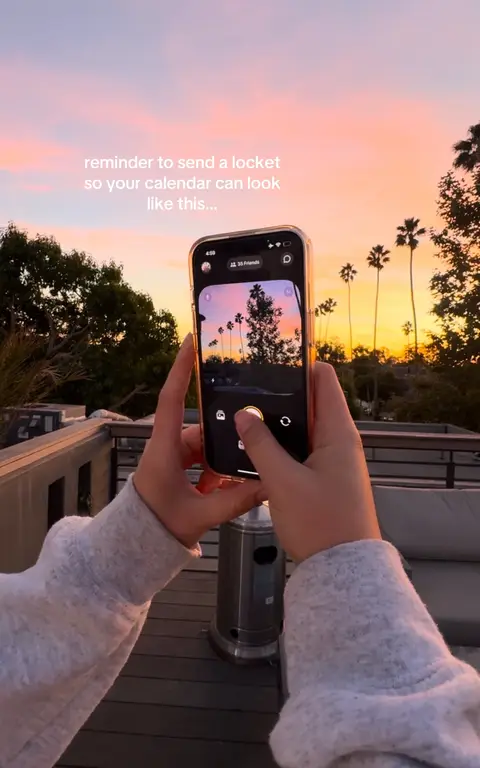 A person is holding a phone, using the back camera to photograph a sunset. The phone's screen displays a nearly identical image of the sunset. The background presents a vibrant sunset with palm trees and a dark silhouette of a building.